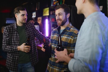 Three young men in casual clothes are smiling, holding bottles of beer while standing near bar counter in pub