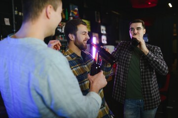 Three young men in casual clothes are smiling, holding bottles of beer while standing near bar counter in pub