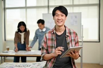 A smart Asian male website developer stands in the office with a digital tablet in his hand