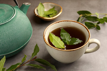 Cup of freshly brewed tea, teapot and bay leaves on grey table, closeup