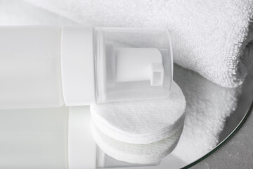 Bottle of face cleansing product, towel and cotton pads on mirror, closeup