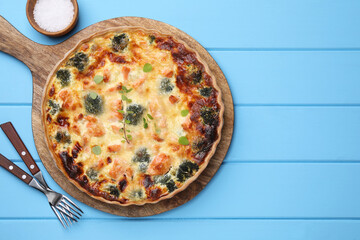 Delicious homemade salmon quiche with broccoli, forks and salt on light blue wooden table, flat lay. Space for text