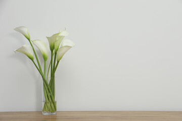 Beautiful calla lily flowers in glass vase on wooden table near white wall. Space for text - obrazy, fototapety, plakaty