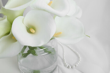 Fototapeta na wymiar Beautiful calla lily flowers in glass vase on white cloth, closeup