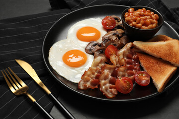 Plate with fried eggs, mushrooms, beans, bacon, tomatoes and toasted bread on black table, closeup. Traditional English breakfast