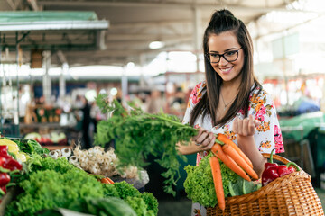 A girl in a white dress gracefully adds fresh carrots to her wooden basket, carefully selecting the vibrant orange vegetables at the market