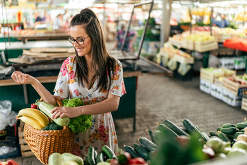 A girl in a white dress carefully chooses an assortment of fresh vegetables, her wooden basket overflowing with a vibrant mix of fruits and vegetables.