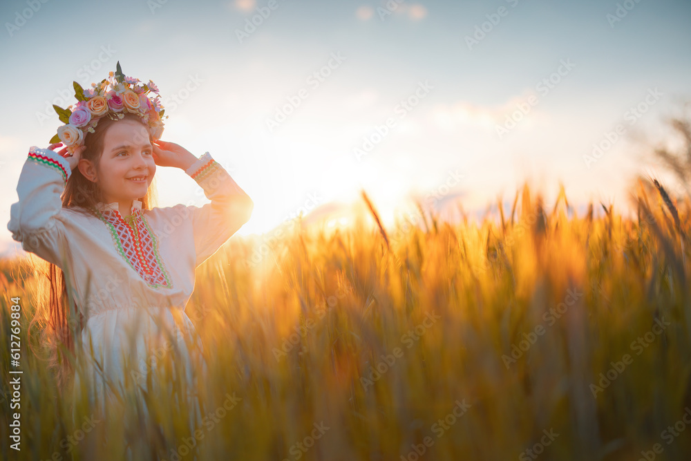 Wall mural Beautiful young girl with flower chaplet, ethnic folklore dress with traditional Bulgarian embroidery during sunset on a wheat agricultural field