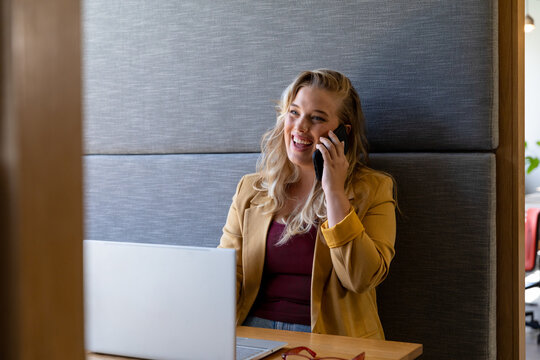 Happy plus size caucasian casual businesswoman using laptop and talking on phone at desk