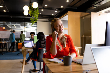 Happy plus size caucasian casual businesswoman using laptop and talking on smartphone at desk