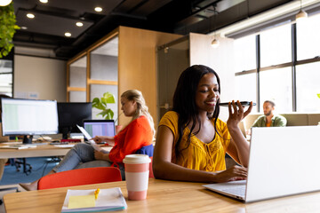 Happy african american casual businesswoman at desk talking on smartphone using laptop in office