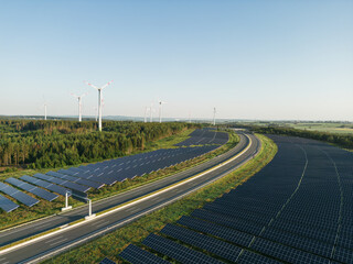 Autobahn zwischen Photovoltaik-Anlagen mit Windrädern im Hintergrund in grüner Natur bei Sonnenschein