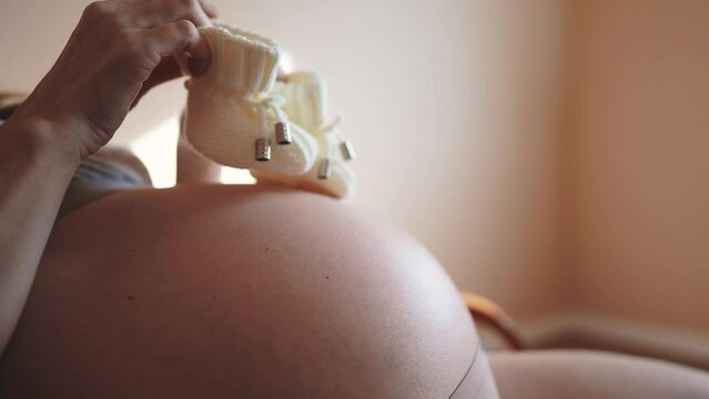 pregnant woman with belly. woman with big bare a belly holding booties baby shoes close-up. health pregnancy motherhood procreation concept. girl indoor preparing for motherhood