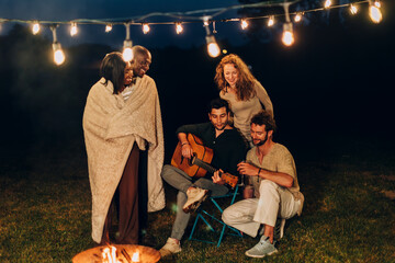 friends sing with guitar around campfire at summer garden birthday party