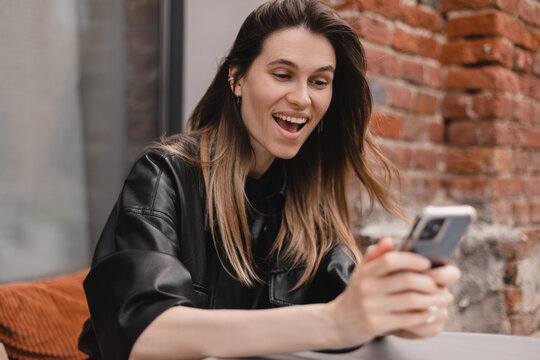 Portrait of happy female dressed casually holding mobile phone, typing messages, communicating with friends via social networks, using high Internet connection at cafe. Woman make video call, wow.