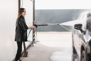 Woman cleaning auto with high pressure water jet at a self-service car wash. Woman wear black...