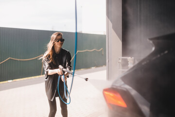 Woman cleaning auto with high pressure water jet at a self-service car wash. Woman wear black...