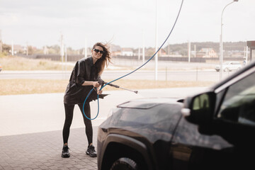 Woman cleaning auto with high pressure water jet at a self-service car wash. Woman wear black...