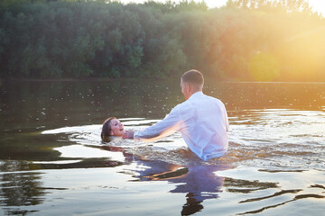 A beautiful adult couple has fun in nature in the water in a river or lake in the summer evening at sunset. A guy and a girl swim and relax outdoors in clothes in white shirts and jeans