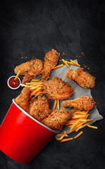 fried chicken bucket dropped on a table, with fries, and crispy golden pieces of chicken meal, ingredients are scattered on the surface of the black table with red sauce perfect poster