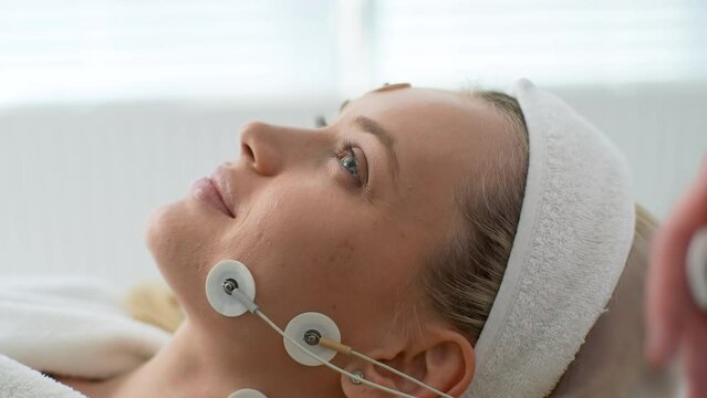 Close-up face of pretty young woman patient preparing for myostimulation procedure. Beautician putting electrodes of myostimulator during bioresonance therapy in medical clinic, slow motion.
