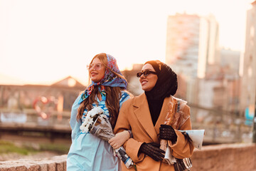 Couple woman one wearing a hijab and a modern yet traditional dress, and the other in a blue dress and scarf, walking together through the city at sunset. One carries a bouquet and bread, while the
