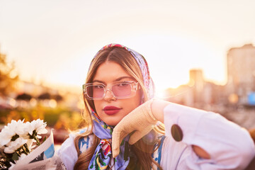 Obraz premium Charm of a woman adorned in an exquisite blue traditional dress, carrying a blue handbag and a bouquet of flowers, gracefully strolling through the city at sunset, creating a mesmerizing scene of