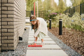 A little girl with a brush cleans a path on the street in the courtyard
