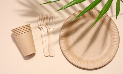 Brown paper plate, stack of glasses and wooden fork, disposable tableware on beige background, top view