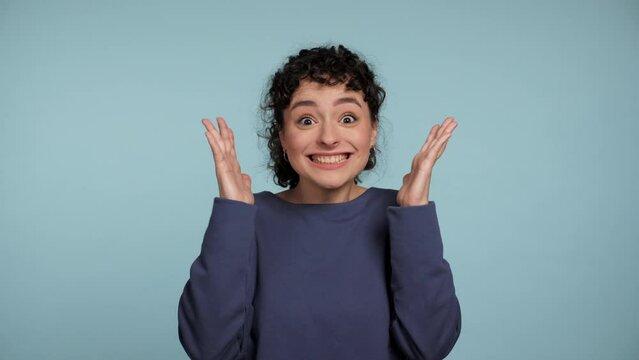 Close up happy young curly woman in blue sweater rejoices waving hands looking camera. Portrait smiling positive female standing on isolated light blue background. Gesture of explosion near temples