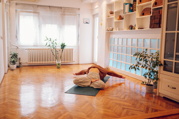 Girl lying on a mat and bending backwards while stretching in a cozy studio.