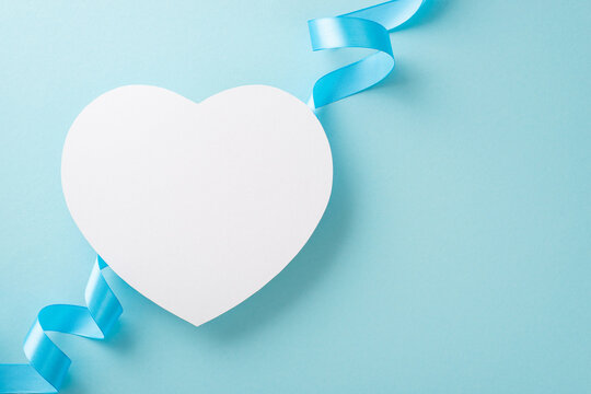 Top view photo of white empty heart-shaped card with a blue ribbon on a light background with copyspace
