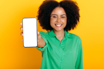 Defocused brazilian or hispanic woman with glasses, in casual shirt, shows cellphone with empty white mockup screen for presentation or advertising, smiles at camera, isolated yellow background