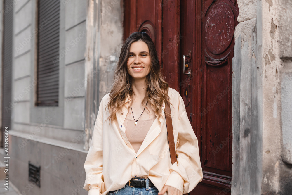 Poster Outdoor portrait of beautiful blonde curly woman standing on step next to old door, old money fashion style. Girl wear yellow shirt, beige top and brown bag. Tourist woman walking outdoor.