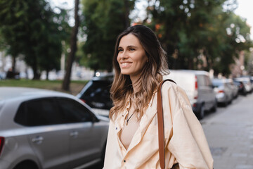 Fototapeta premium Blonde coquette woman look at side, flirting. Woman wear yellow shirt, beige top, brown bag, walking outdoors near cars. Happy tourist traveler girl.