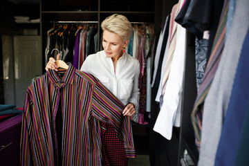 Mature Woman stylist organizes or chooses clothes outfit and tries on in large wardrobe closet at...