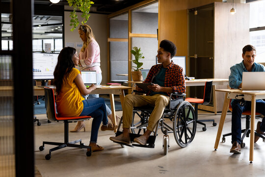 Diverse female and male colleague in wheelchair in discussion using tablet at desk in casual office