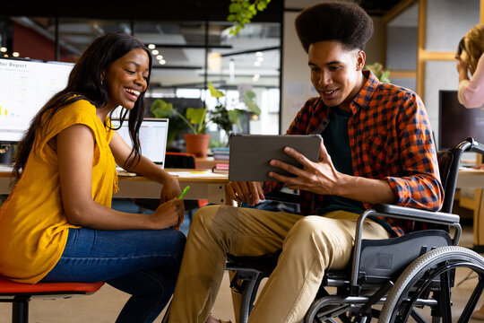 Happy diverse female and male colleague in wheelchair using tablet at desk in casual office