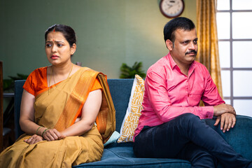 Indian angry middle aged couple sitting on sofa in opposite direction at home - concept of...