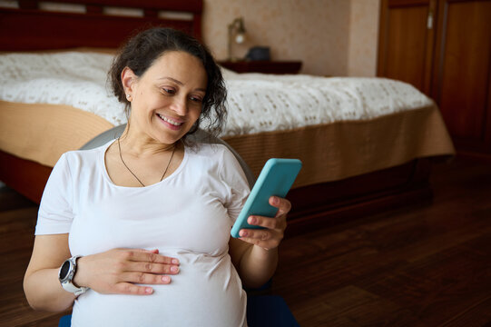Smiling pregnant woman keeps her hand on her big belly, counts the kicks of the baby and checks a mobile application for pregnant women on her smartphone while relaxing, sitting on the floor at home