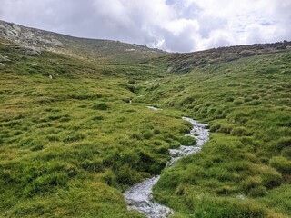 Paisaje de alta montaña con un arroyo atravesando un prado verde.