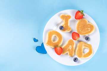 Father's day holiday idea for a cute surprise gift. Child preparing breakfast for dad, creatively decorated pancakes with the inscription I love Dad. Top view copy space, blue background