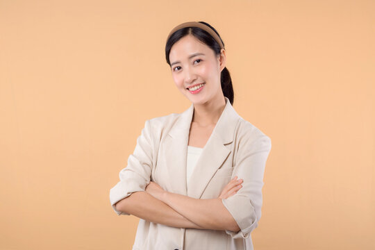 Studio Portrait Of Happy Successful Confident Young Asian Business Woman. Beautiful Young Lady In White Jacket Smiling At Camera Standing Isolated On Solid Beige Colour Copyspace Background