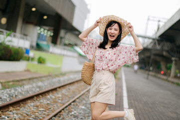 young asian woman traveler with weaving basket happy smiling looking to a camera beside train railway. Journey trip lifestyle, world travel explorer or Asia summer tourism concept.