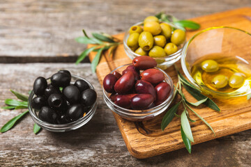 Green, black and red olives on a brown wooden background. Various types of olives in bowls and olive oil with fresh olive leaves. Copy space. Place for text. Mediterranean food. Vegan.