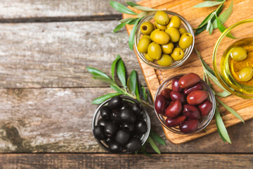 Green, black and red olives on a brown wooden background. Various types of olives in bowls and olive oil with fresh olive leaves. Copy space. Place for text. Mediterranean food. Vegan.