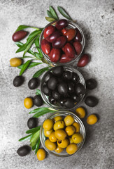 Green, black and red olives on a textured marble background. Various types of olives in bowls and olive oil with fresh olive leaves. Copy space. Place for text. Mediterranean food. Vegan.