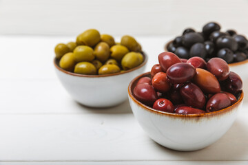 Green, black and red olives on a white wooden background. Various types of olives in bowls and olive oil with fresh olive leaves. Copy space. Place for text. Mediterranean food. Vegan.