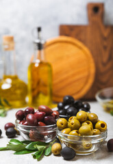 Green, black and red olives on a textured marble background. Various types of olives in bowls and olive oil with fresh olive leaves. Copy space. Place for text. Mediterranean food. Vegan.