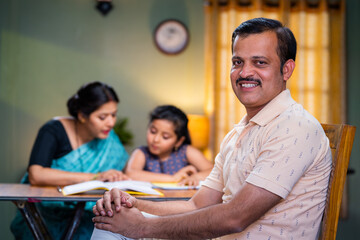 Happy indian father looking at camera in front of mother helping daughter for reading at home - concept of responsibility, successful family and patriarchal society.